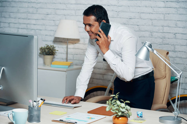 Homme d'affaires indien regardant moniteur d'ordinateur tout en répondant à un appel téléphonique sur le lieu de travail
 - Photo, image