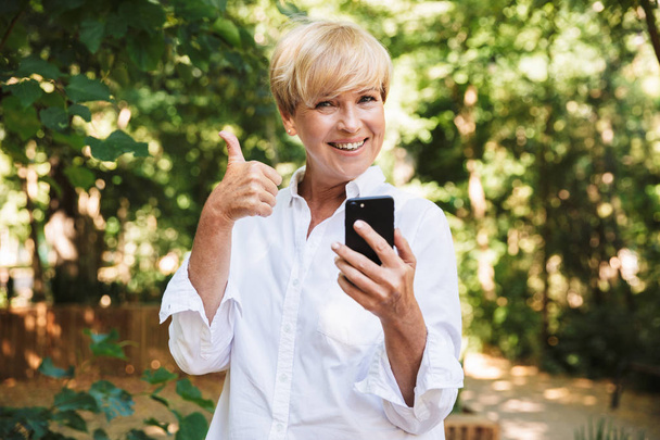 Happy mature woman using mobile phone and showing thumbs up while resting at the park - Foto, Imagen