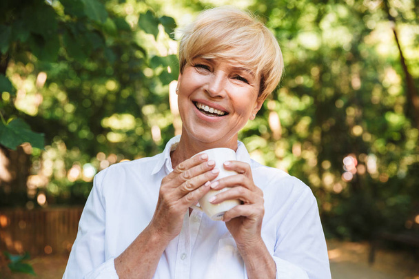 Happy mature woman holding cup of coffee while resting at the park - Fotoğraf, Görsel