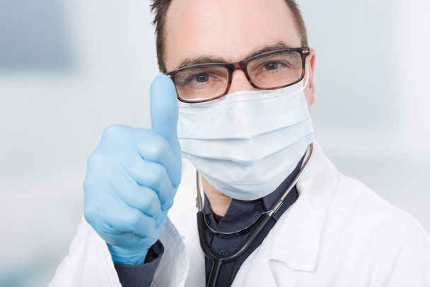 ο γιατρός με ιατρική μάσκα και γάντια υγιεινής παρουσιάζει αντίχειρα - Φωτογραφία, εικόνα