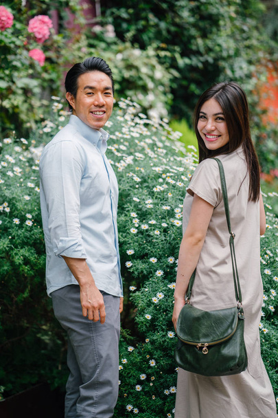 A young and attractive Asian couple stop to smell the flowers in a park during the day.  - Photo, Image