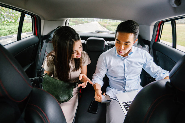 Two young Asian business people sit on the back seat of a car and are driven to their destination on a ride they booked via a ride hailing app. - Fotografie, Obrázek