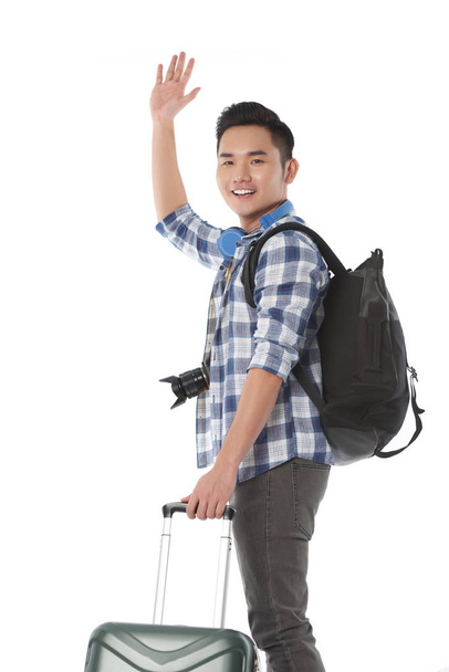 Joyful Asian traveler man holding handle of suitcase and waving hand to camera, isolated on white background - Photo, Image