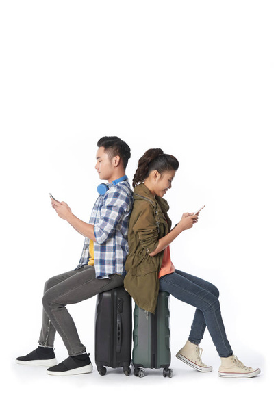 asiatisches Paar sitzt auf Koffern und surft auf Smartphones, isoliert auf weißem Hintergrund - Foto, Bild