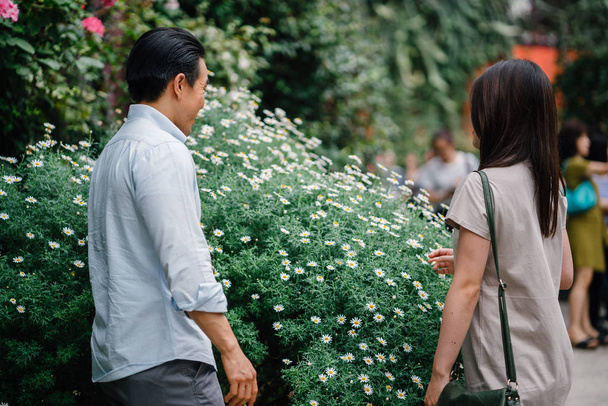 A young and attractive Asian couple stop to smell the flowers in a park during the day.  - Foto, Imagen
