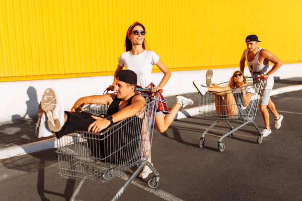 Friends ride on carts, near the supermarket, a great pastime, a group of young people rides on carts, couples in love have fun in Sunny weather - Zdjęcie, obraz