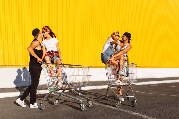 Kissing lovers sitting on carts sitting on the cart, near the supermarket, a great pastime, a group of young people rides on carts, couples in love have fun in Sunny weather - 写真・画像