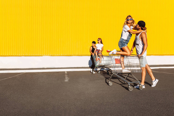 Kissing lovers sitting on carts sitting on the cart, near the supermarket, a great pastime, a group of young people rides on carts, couples in love have fun in Sunny weather - 写真・画像