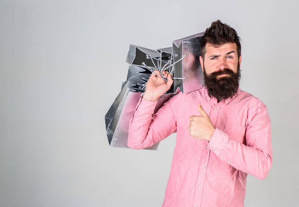 Guy shopping and shows thumbs up gesture. Hipster on confident face recommends to buy. Black friday concept. Man with beard and mustache carries bunch of shopping bags, grey background - Φωτογραφία, εικόνα