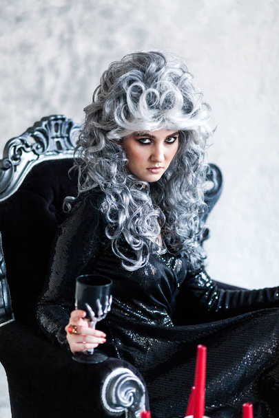 Halloween. Portrait of a gorgeous young glamour beautiful woman vampire baroque aristocrat witch in black dress with gray curly hair with a black glass in the ancient castle. - Photo, Image