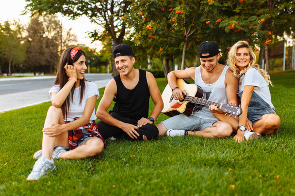 Company of friends having fun at sunset sitting on the grass, playing guitar, good summer mood - Foto, imagen