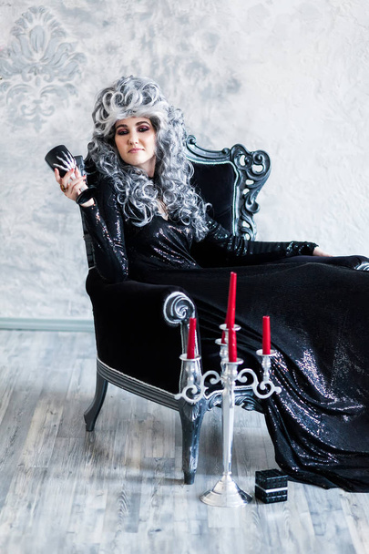 Halloween. Portrait of a gorgeous young glamour beautiful woman vampire baroque aristocrat witch in black dress with gray curly hair with a black glass in the ancient castle. - Photo, Image