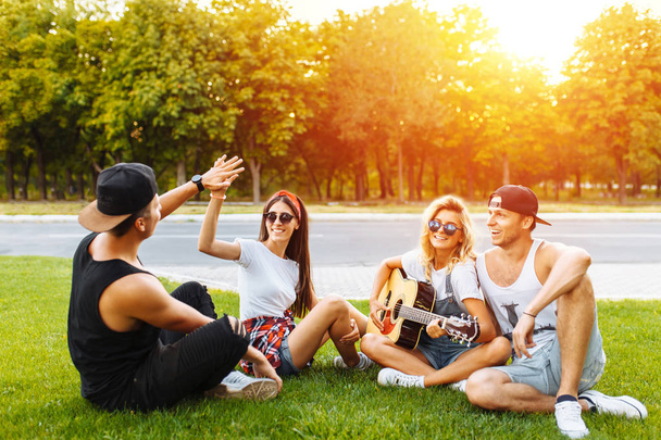 Company of friends having fun at sunset sitting on the grass, playing guitar, good summer mood - Fotografie, Obrázek