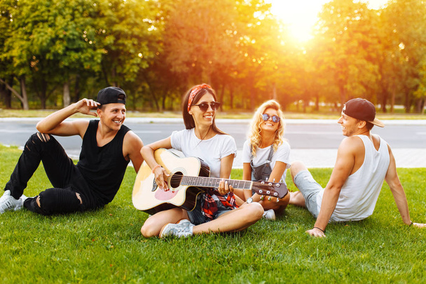 Company of friends having fun at sunset sitting on the grass, playing guitar, good summer mood - Foto, afbeelding