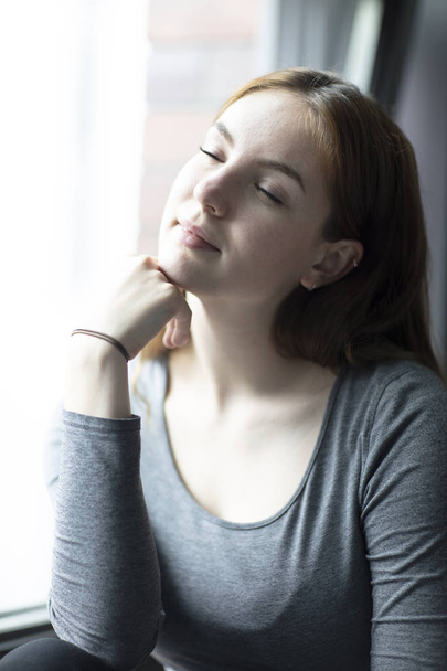 Portret młodej kobiety z wyrazem zachwytu z zamkniętymi oczami siedząc przez okno. Średni strzał. Selektywny fokus.  - Zdjęcie, obraz
