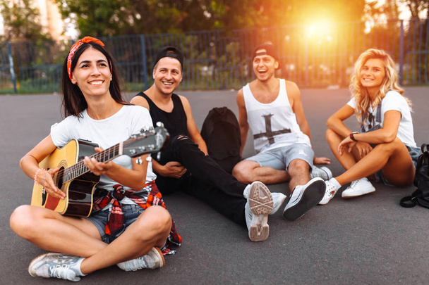 A group of friends having fun at sunset on the asphalt in a skate Park, playing guitar, good summer mood - Foto, afbeelding