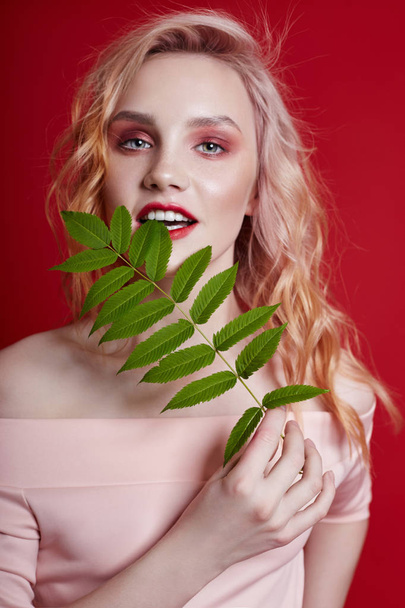 Sexy fashion blonde with bright red makeup and branch fern on bright red background. Girl with a perfect figure. Professional make-up, skin care, natural cosmetics for face, beautiful eyes - Foto, afbeelding