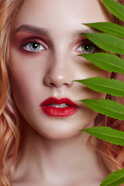 Sexy fashion blonde with bright red makeup and branch fern on bright red background. Girl with a perfect figure. Professional make-up, skin care, natural cosmetics for face, beautiful eyes - Photo, image
