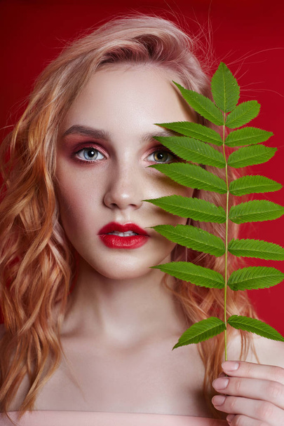 Sexy fashion blonde with bright red makeup and branch fern on bright red background. Girl with a perfect figure. Professional make-up, skin care, natural cosmetics for face, beautiful eyes - Фото, зображення