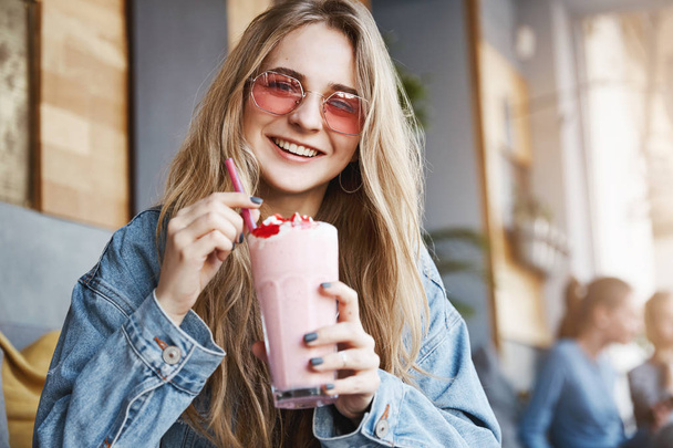 Wanna share cocktail with me. Joyful and carefree friendly-looking caucasian woman in trendy pink sunglasses, holding pink and tasty drink, smiling happily while having funny conversation - Fotografie, Obrázek