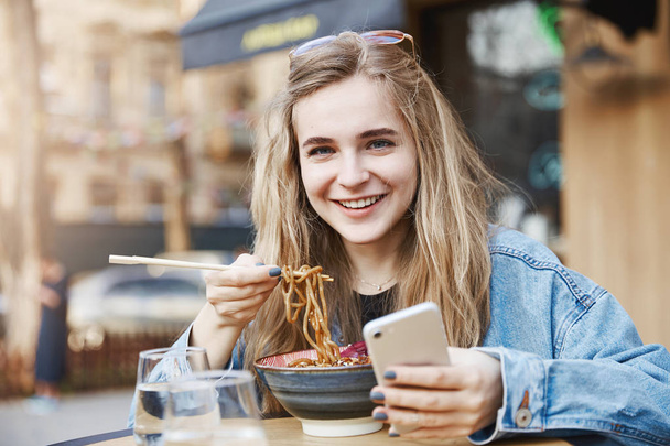 Stylish teenage girl cannot leave smartphone even while eating. Portrait of pleased and happy good-looking woman in trendy outfit and glasses, holding phone and chopsticks, eating asian food on street - Photo, image