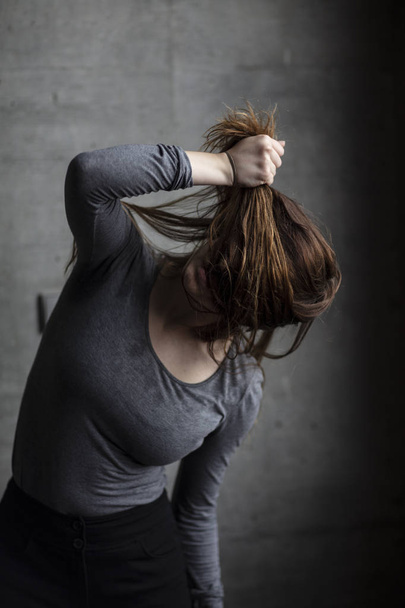 Female model posing behind a grey concrete wall pulling her hair and obscuring her face. Medium shot.  - Foto, afbeelding
