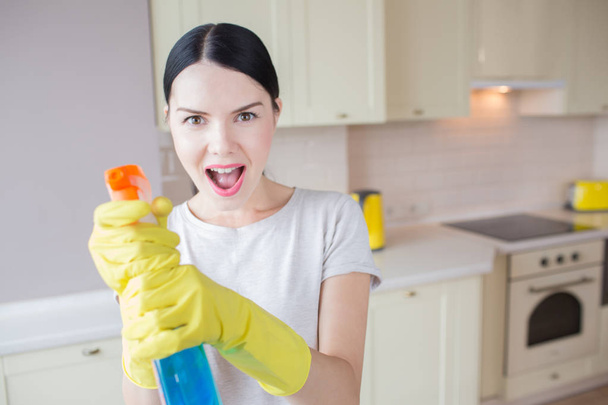 A picture of excited woman stands and holds blue spray in hands. She aims it on camera. Girl stands in kitchen. - Foto, imagen
