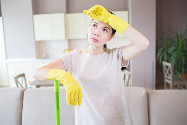 Tired and exhausted woman stands in the middle of room and holds left hand on forehead. She wears yellow gloves. Girl is leaning to green stick with another hand. - Photo, image