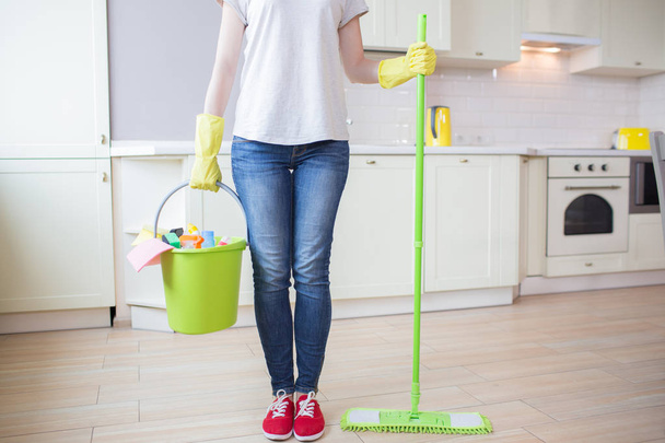 Cut view of woman stands in kitchen and holds stick with mop in one hand and bucket with cleaning equipment in the other one. Girl wears yellow gloves. - Photo, Image