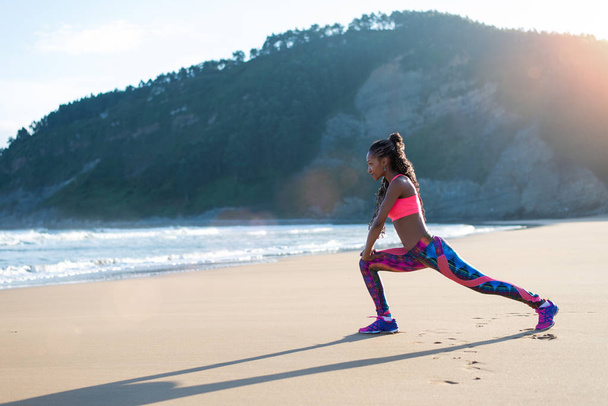 Фитнес-женщина растягивает ноги или делает выпады на пляже. Черная спортсменка делает расслабляющие упражнения в сторону моря
. - Фото, изображение