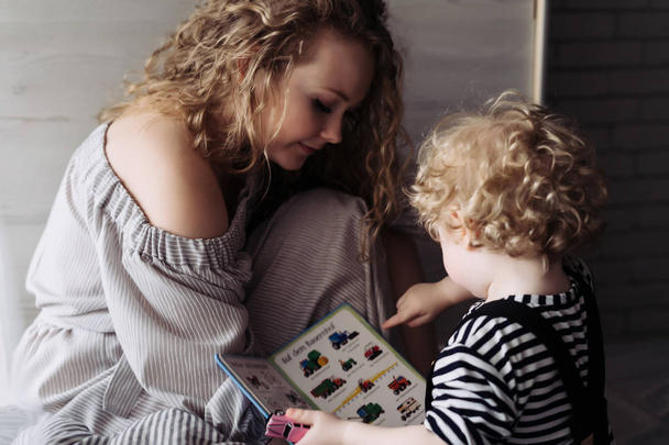 curly young mother in a dress spends time with her fair-haired son, read books - Foto, Bild