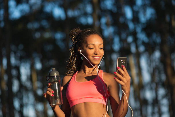 Fitness brazilian sportswoman taking a break for drinking water and listening music on smart phone during outdoor workout. Black female athlete wearing earphones. - Photo, Image