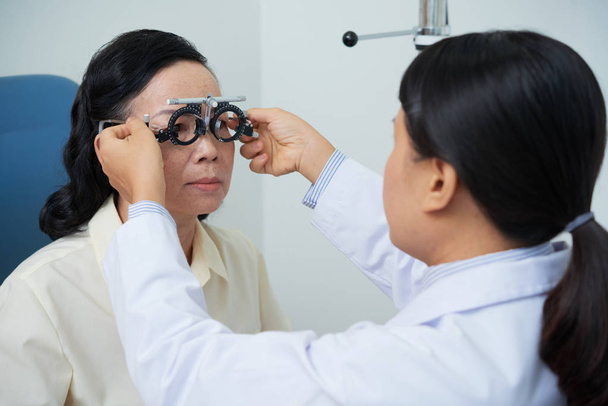 Professional vision specialist taking care of Asian patient in cabinet of hospital and testing eyesight with trial lens eyeglasses - Photo, image