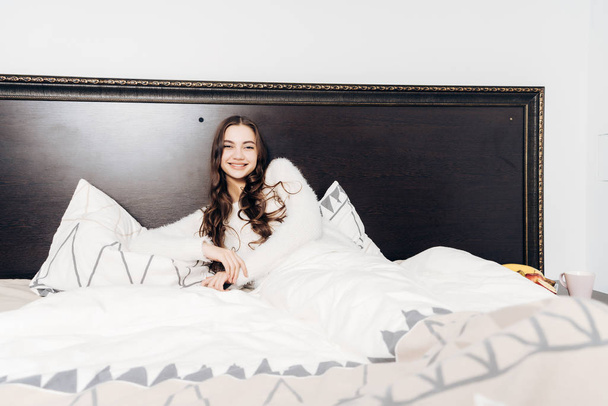 beautiful sleepy girl lies under a white blanket in the early morning, does not want to get up, smiles - Fotografie, Obrázek