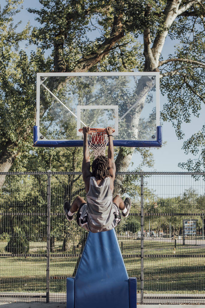 Back view of an African basketball player dunking a basketball and hanging on a basketball rim. - Fotografie, Obrázek