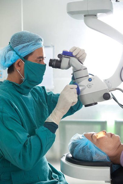 Aasialainen lääkäri mies kirurgin univormussa tutkii kypsän potilaan silmiä leikkausmikroskoopilla
 - Valokuva, kuva