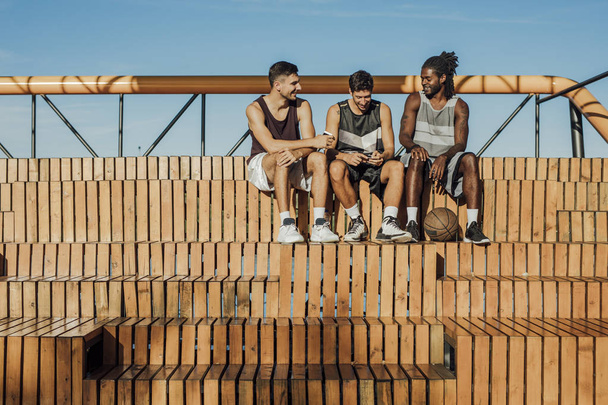 Three handsome friends and basketball players having fun while using their mobile phones and sitting on the outdoor basketball bleachers. - Zdjęcie, obraz