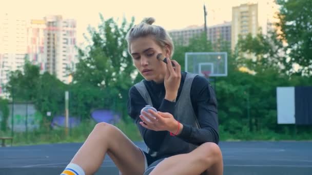 Attractive female basketball player sitting on court and doing make up, park and buildings in background - Metraje, vídeo
