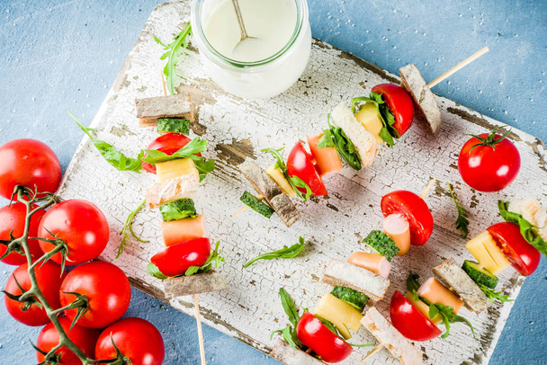 Healthy summer snack idea, salad sandwiches kebab on skewers, with bread slices, tomato, greens, cucumber, cheese, sausages, with yoghurt dipping, on light concrete background copy space top view - Photo, Image