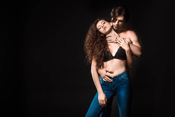 man hugging seductive girlfriend in black bra and jeans isolated on black - Photo, Image