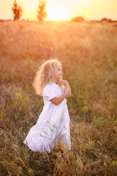 Αξιολάτρευτο ευτυχισμένο κοριτσάκι με μια σγουρά ξανθά μαλλιά, φορώντας ένα λευκό φόρεμα, που στέκεται στο ηλιόλουστο ηλιοβασίλεμα πεδίο ανάμεσα σε άγρια χλόη και λουλούδια, χαμογελώντας - Φωτογραφία, εικόνα