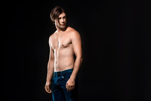 portrait of pensive shirtless man in jeans looking away isolated on black - Fotó, kép