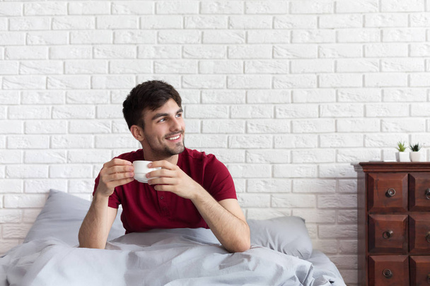 Όμορφος άντρας πίνει καφέ, ενώ κάθεται στο κρεβάτι - Φωτογραφία, εικόνα