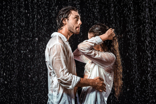 Seitenansicht eines Paares in weißen Hemden, das unter Regen isoliert auf schwarz steht - Foto, Bild