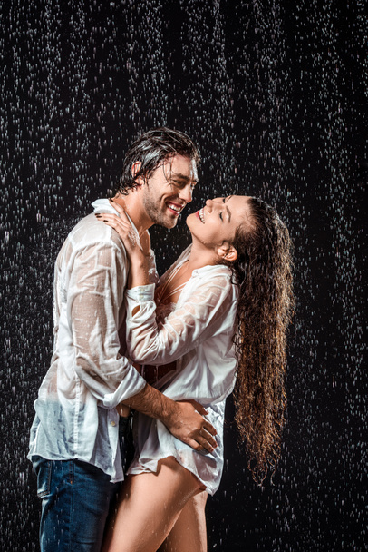 vista lateral de la pareja sonriente abrazándose mientras está de pie bajo la lluvia aislado en negro
 - Foto, imagen