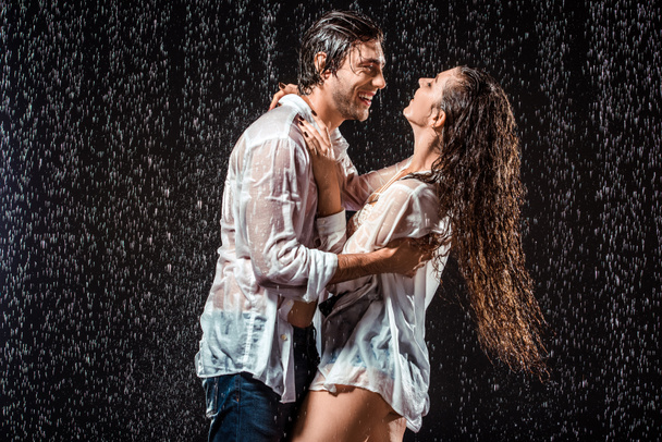 happy wet couple in white shirts standing under rain isolated on black - Photo, Image