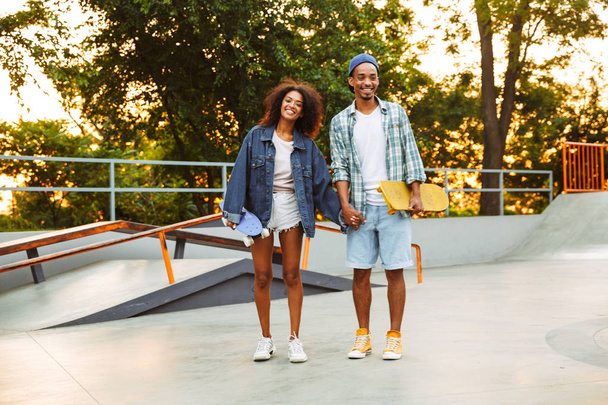 Portrait of a cheerful young african couple with skateboards standing together at the skate park - Photo, Image