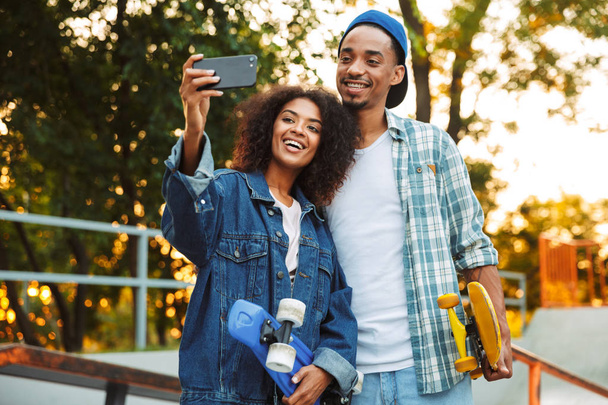 Retrato de una joven pareja africana sonriente con monopatines tomando una selfie juntos en el parque de skate
 - Foto, Imagen