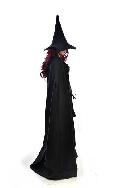 Ganzkörperporträt eines rothaarigen Mädchens, das einen langen schwarzen Mantel, einen spitzen Hut und ein Hexenkostüm trägt. Stehende Pose, isoliert auf weißem Studiohintergrund. - Foto, Bild