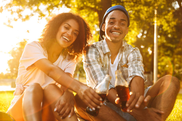 Image of cute cheerful young african loving couple sitting outdoors in park drinking soda. - Photo, Image